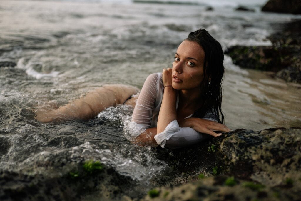 Corps complet d'une jeune femme mince en chemise blanche et bikini noir, allongée les bras croisés sur une côte sablonneuse tandis que l'eau de mer agite et couvre le corps de la femme, chirurgie de féminisation du corps