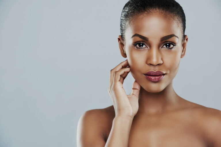 Una mujer con los hombros descubiertos y los dedos cerca de la mandíbula, mirando directamente a la cámara con una expresión seria, sobre un fondo liso.