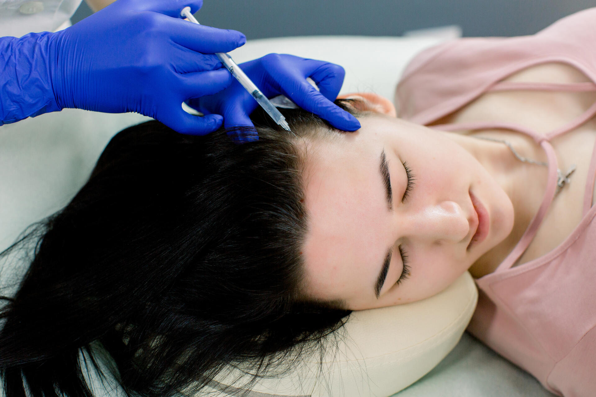 A beautician performing hair mesotherapy on a client. The client is lying down with eyes closed while the beautician, wearing blue gloves, uses a syringe to inject a solution into the client's scalp.