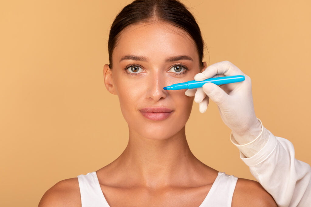Notion de rhinoplastie. Médecin faisant des marques sur le visage du patient, jeune femme caucasienne en consultation chez un chirurgien, debout sur fond beige. Concept de chirurgie plastique du visage
