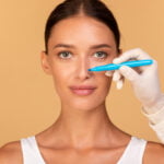 Notion de rhinoplastie. Médecin faisant des marques sur le visage du patient, jeune femme caucasienne en consultation chez un chirurgien, debout sur fond beige. Concept de chirurgie plastique du visage