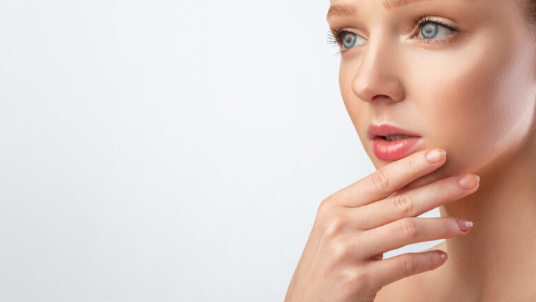 Mujer con maquillaje perfecto posando tocando la barbilla, fondo gris, recortada, cuidado de la piel facial. Mujer bastante joven con maquillaje perfecto posando tocando la barbilla y mirando a un lado el espacio libre para texto sobre fondo gris de estudio. Panorámica, recortada