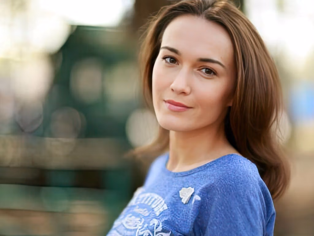 A woman with long brown hair wearing a blue shirt with a floral pattern sitting on a bench in an outdoor setting.