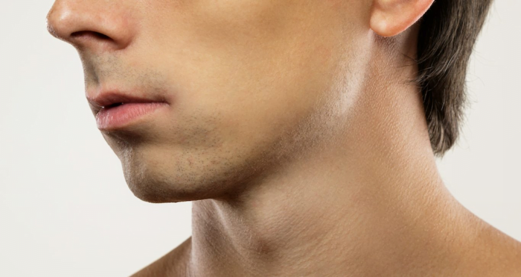 Gros plan de profil du visage d'une personne, mettant l'accent sur la mâchoire et le cou, sur un fond uni. L'image met en valeur le visage rasé de près et la texture lisse de la peau. Les lèvres sont légèrement entrouvertes et l'aile du nez est également visible.