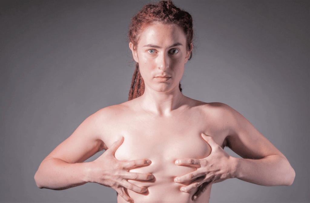 Una persona a torso nudo, con i capelli corti e ricci tirati indietro, che si tiene il petto con entrambe le mani, su uno sfondo grigio semplice.