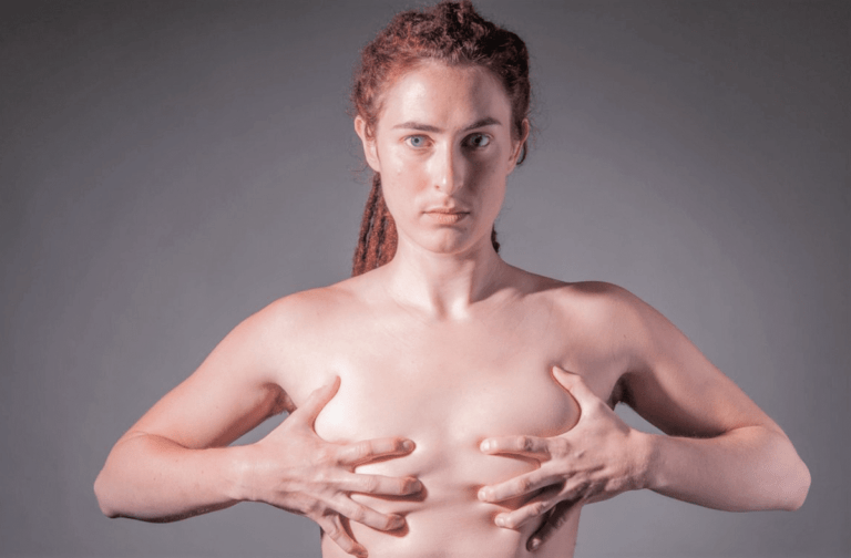 Человек без рубашки, с короткими вьющимися волосами, зачесанными назад, держащийся за грудь обеими руками, на однотонном сером фоне.