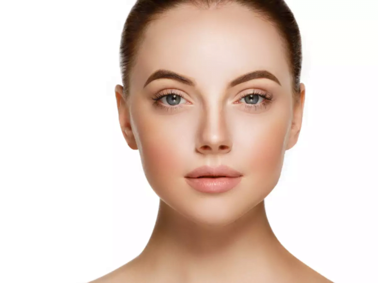 Портрет крупным планом женщины с гладкой кожей, четко очерченными бровями и нейтральным выражением лица на простом белом фоне.