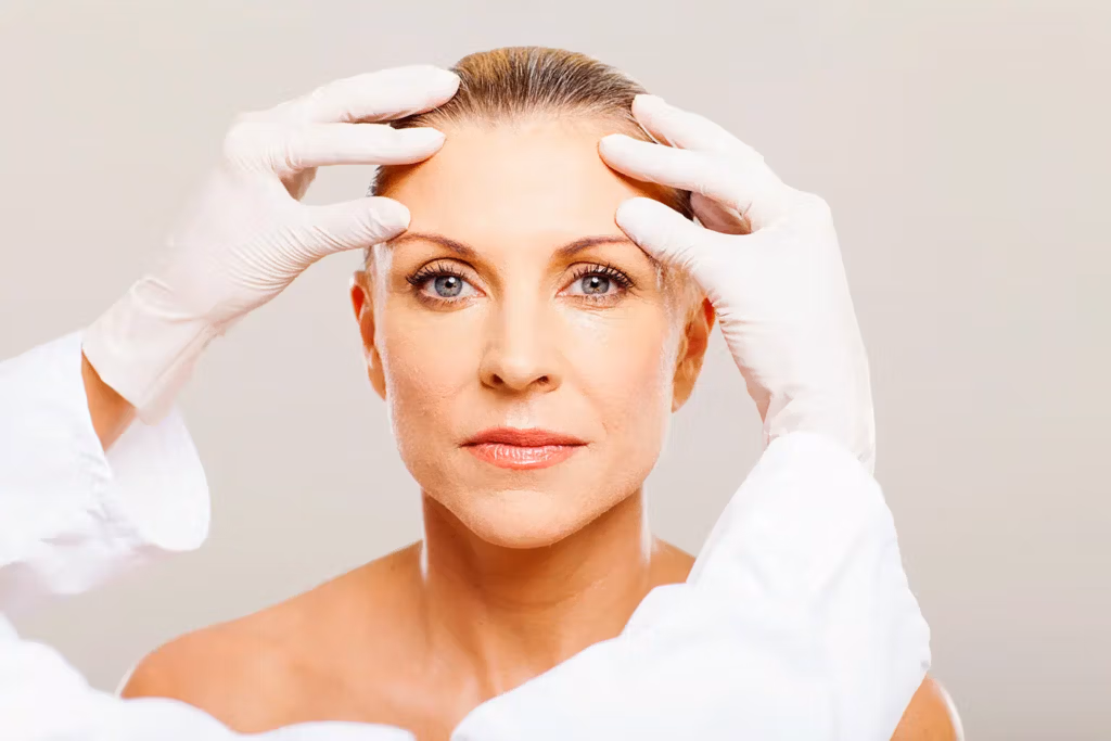 Fotografía en primer plano de una mujer madura sometiéndose a un procedimiento médico. Sus manos enguantadas se tocan suavemente la frente y las sienes. La mujer luce tranquila y concentrada, con la mirada fija al frente. La imagen transmite profesionalismo y atención en un entorno clínico.