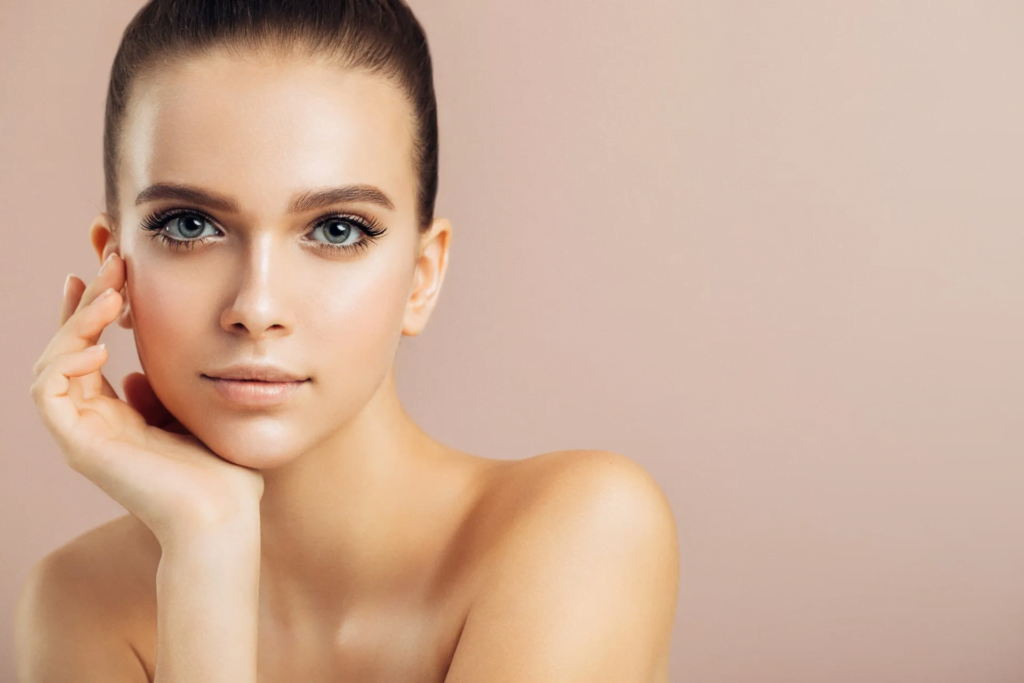 A close-up portrait of a woman with short hair and striking blue eyes, posing with her hand gently touching her face. The background is a soft, neutral tone, emphasizing her natural beauty and minimal makeup.