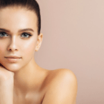 Un ritratto ravvicinato di una donna con i capelli corti e gli occhi azzurri, in posa con una mano che le sfiora delicatamente il viso. Lo sfondo è in tonalità tenui e neutre, che ne enfatizzano la bellezza naturale e il trucco minimale.