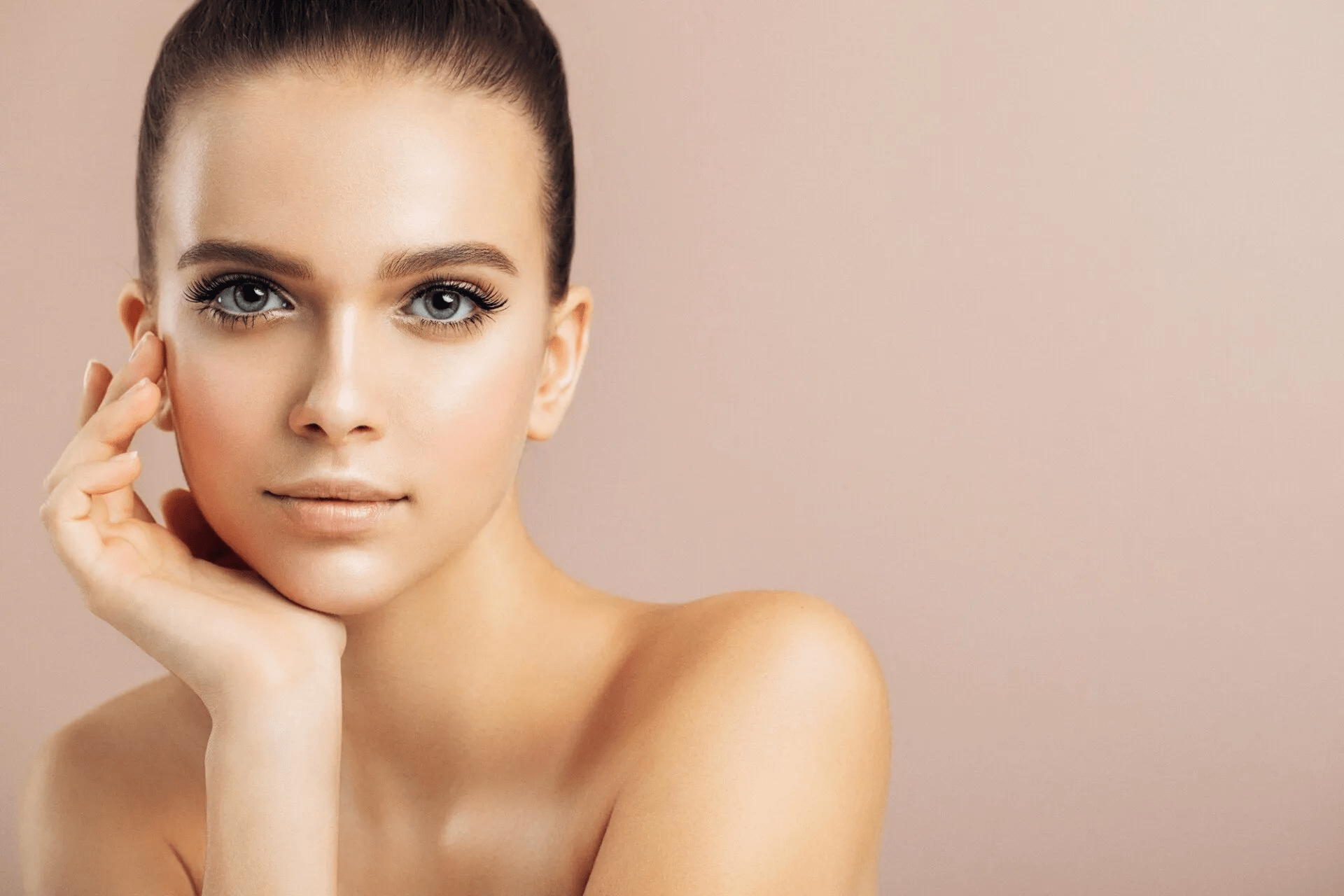 A close-up portrait of a woman with short hair and striking blue eyes, posing with her hand gently touching her face. The background is a soft, neutral tone, emphasizing her natural beauty and minimal makeup.