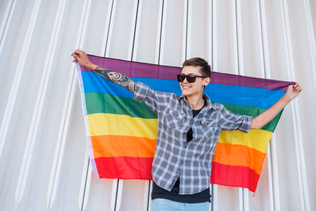Eine Person mit kurzen Haaren und Sonnenbrille, die ein kariertes Hemd und Ohrringe trägt, lächelt und vor einer weißen Wellblechwand eine große Regenbogenflagge hält.