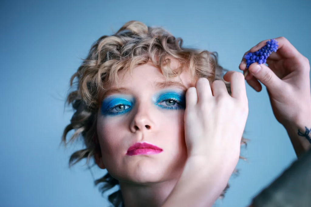 Portrait en gros plan d'une personne aux cheveux bouclés, tenant une grappe de raisin violet près de son visage. Elle porte un fard à paupières bleu et vert vif et un rouge à lèvres rose. Le fond est d'un bleu clair uni.