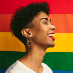 Een persoon met krullend haar en een stralende glimlach, gekleed in een wit shirt en grote oorringen, staat voor een achtergrond met een levendige regenboogvlag. De uitdrukking van de persoon is vrolijk en zelfverzekerd, wat trots en feestvreugde symboliseert.