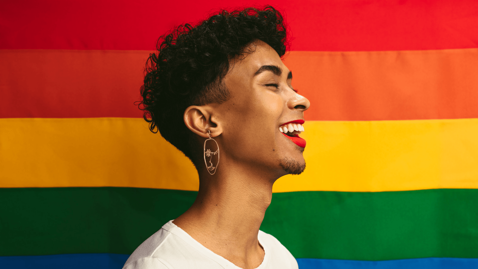A person with curly hair and a bright smile, wearing a white shirt and large hoop earrings, stands in front of a vibrant rainbow flag background. The person's expression is joyful and confident, symbolizing pride and celebration.