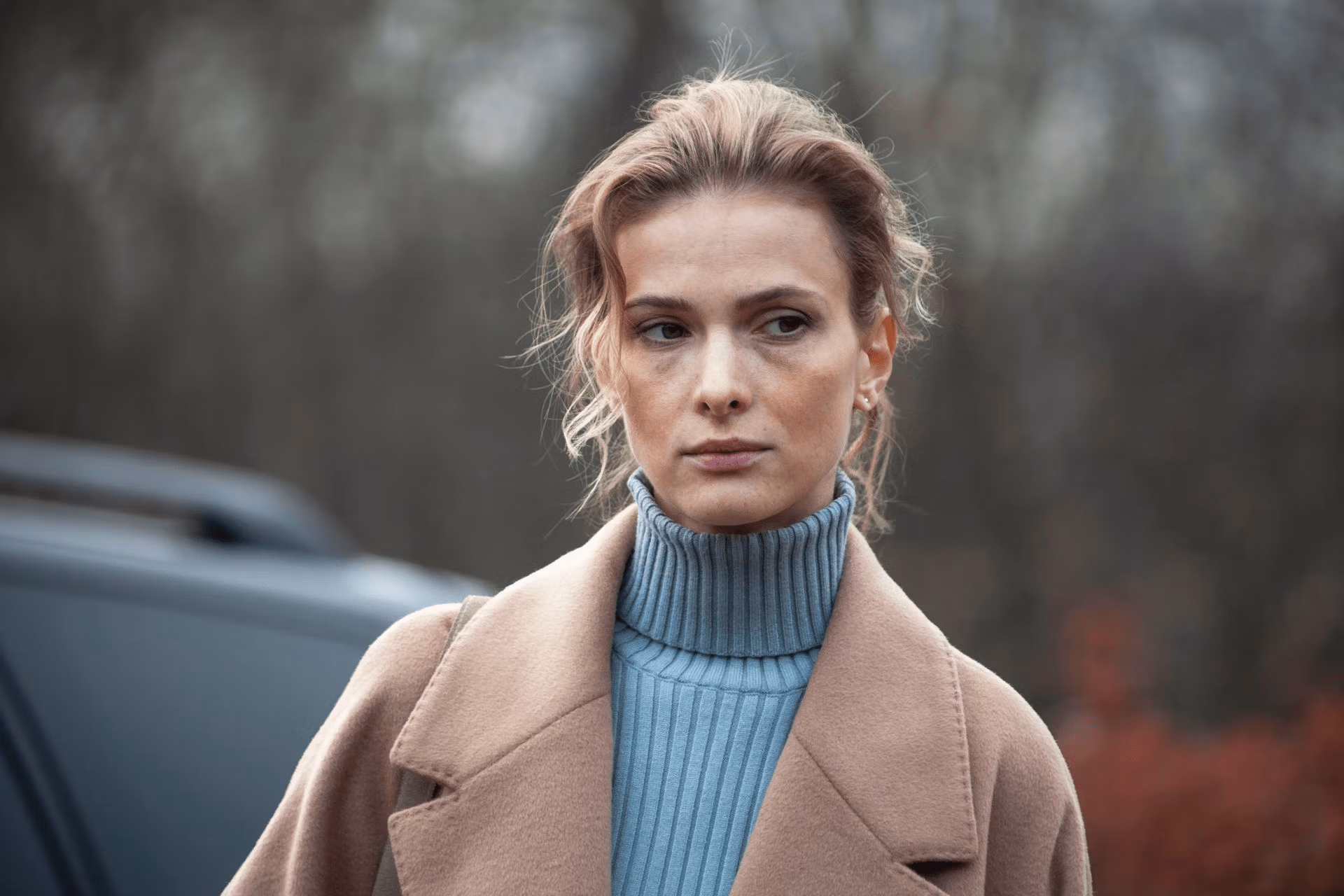 A person standing outdoors, wearing a beige coat over a light blue turtleneck sweater, with a serious expression on their face and hair pulled back.