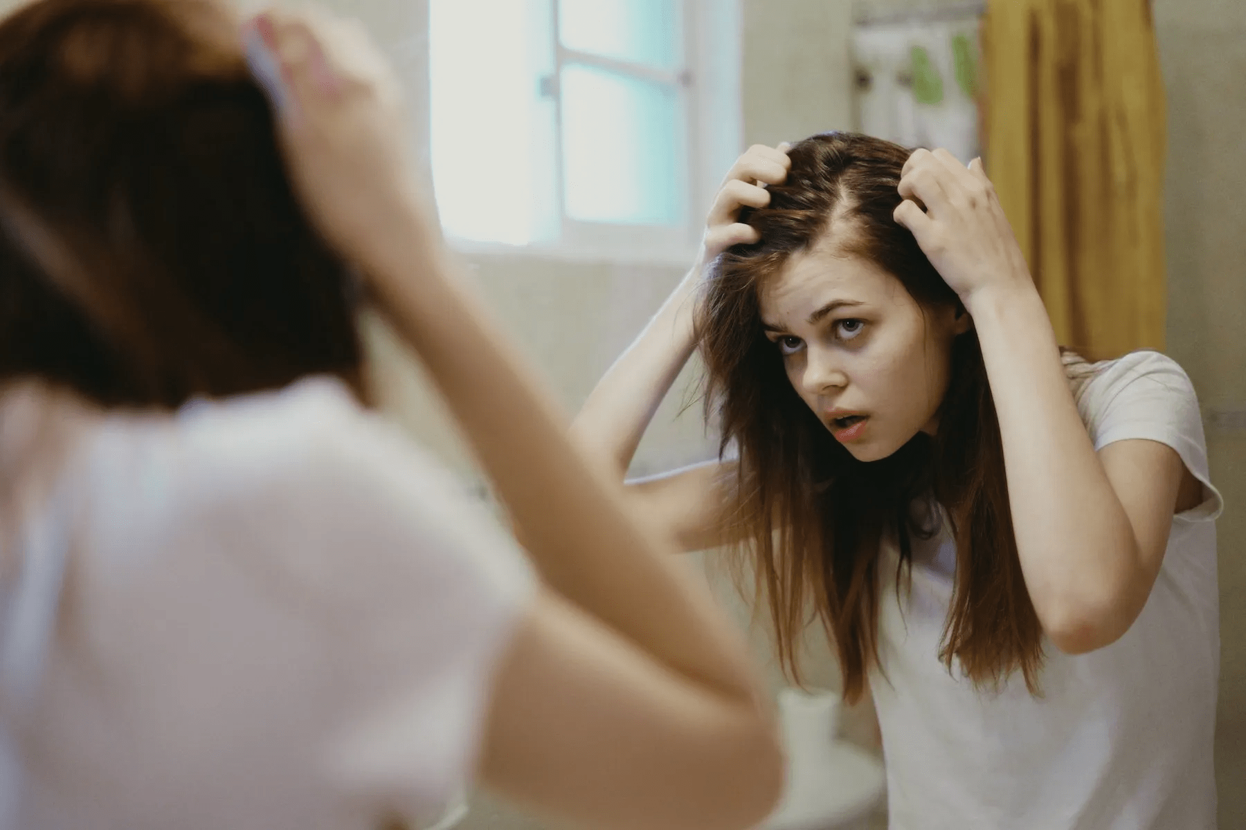 A person with long hair looking at their reflection in a mirror, adjusting their hair with both hands, appearing to be in a bathroom setting.