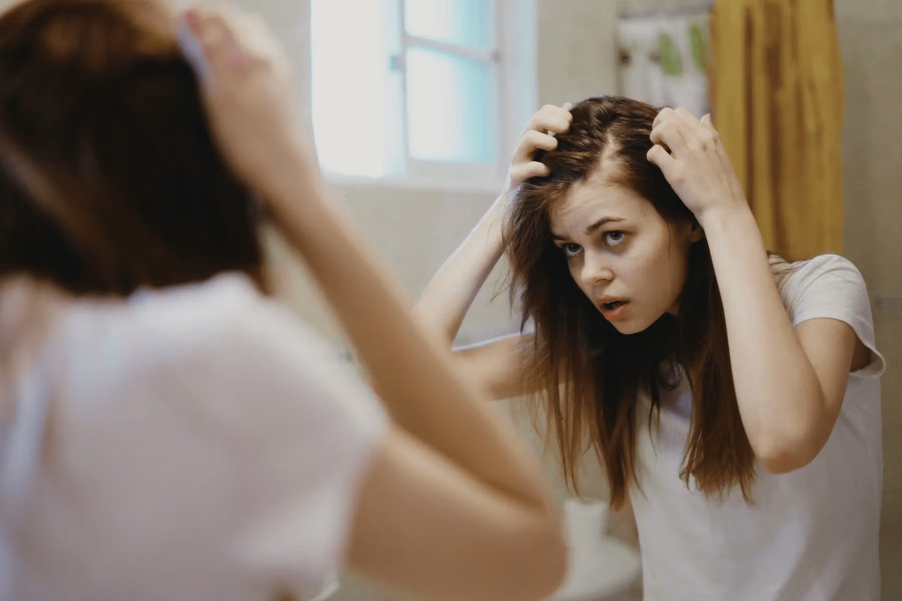 A person with long hair looking at their reflection in a mirror, adjusting their hair with both hands, appearing to be in a bathroom setting.