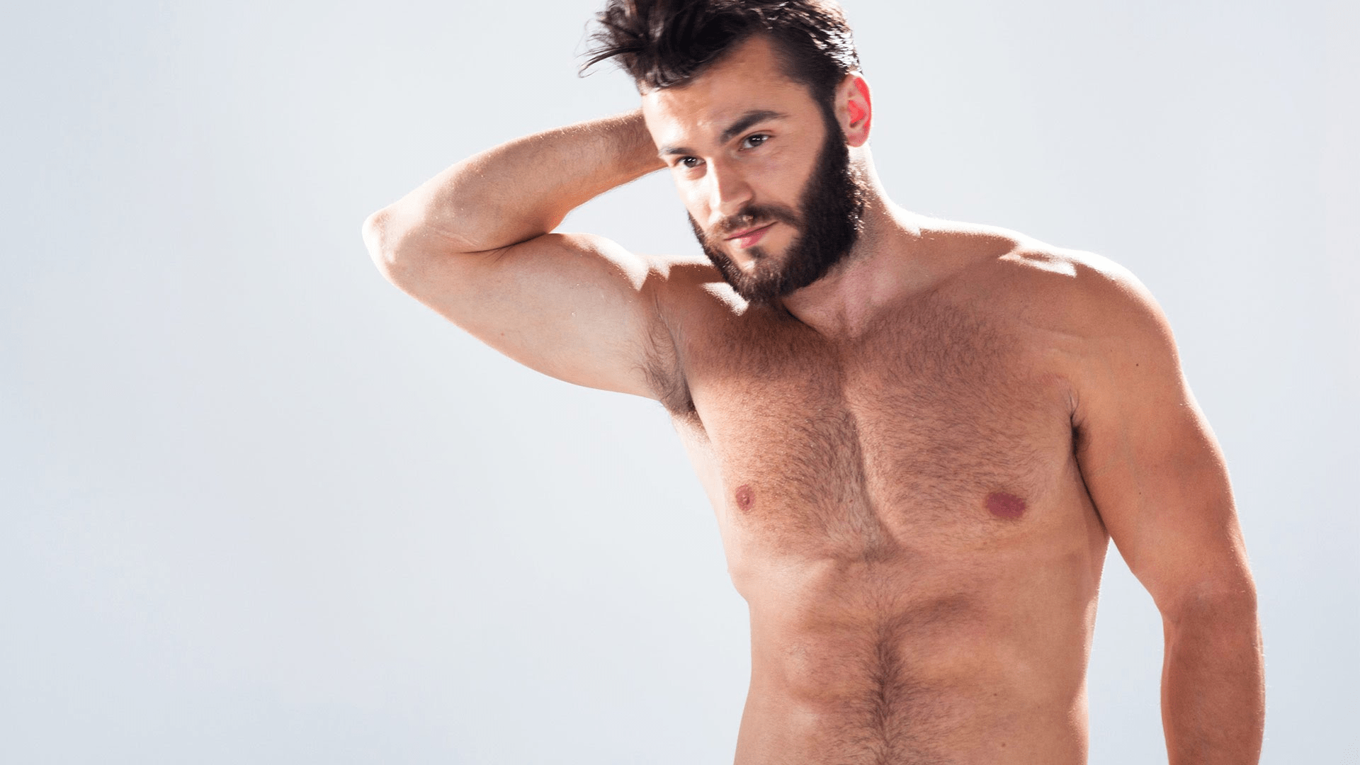 A shirtless man with a beard and short hair, standing with one hand on his head, looking directly at the camera against a plain white background.