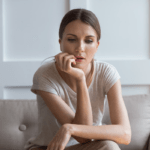 A woman sitting on a beige couch with her hand on her chin, looking thoughtful. The room has white walls and minimal decor, with a single green plant visible in the background.