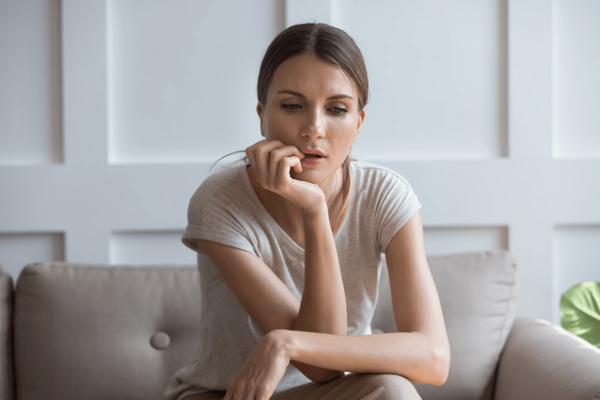 A woman sitting on a beige couch with her hand on her chin, looking thoughtful. The room has white walls and minimal decor, with a single green plant visible in the background.