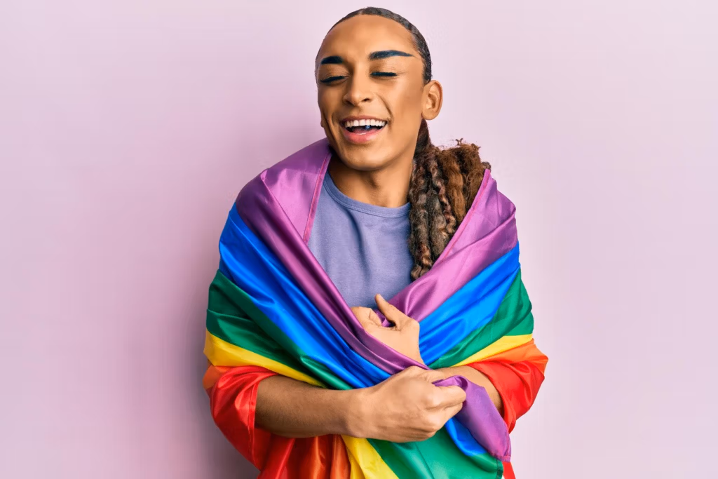 Una persona con expresión alegre, vestida con una camisa gris, envuelta en una vibrante bandera arcoíris, que simboliza el orgullo y el apoyo LGBTQ+.