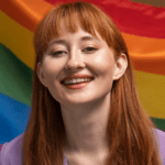 A smiling woman with long red hair and bangs, wearing a purple top, standing in front of a large rainbow flag.