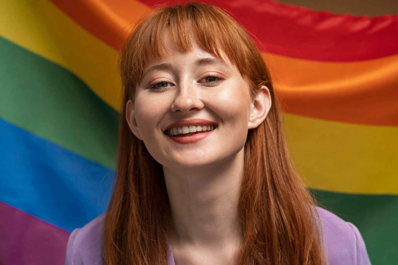 A smiling woman with long red hair and bangs, wearing a purple top, standing in front of a large rainbow flag.