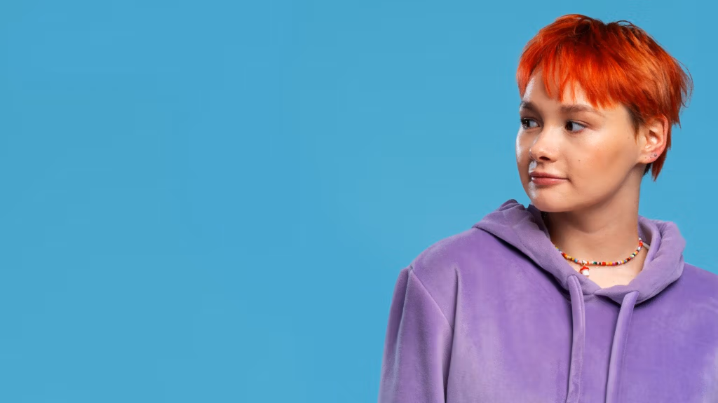 Una persona con el pelo corto de color naranja, que lleva una sudadera con capucha morada y un collar colorido, de pie sobre un fondo azul liso.