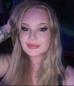 Nahaufnahme einer Person mit langen blonden Haaren, leichtem Make-up und einem leichten Lächeln. Die Person trägt eine Halskette und hat ein Tattoo auf dem linken Arm. Das Bild enthält ein Wasserzeichen für Dr. MFO Facial Surgery & Beauty.