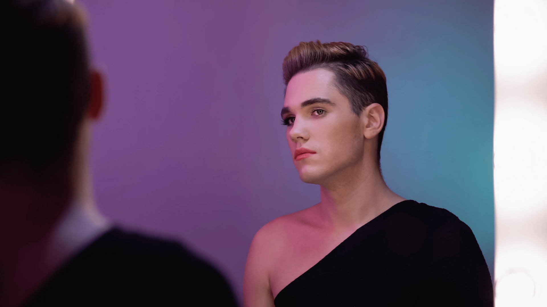 A person with short, styled hair looking to the side, wearing a black off-shoulder top, standing against a gradient purple and blue background.