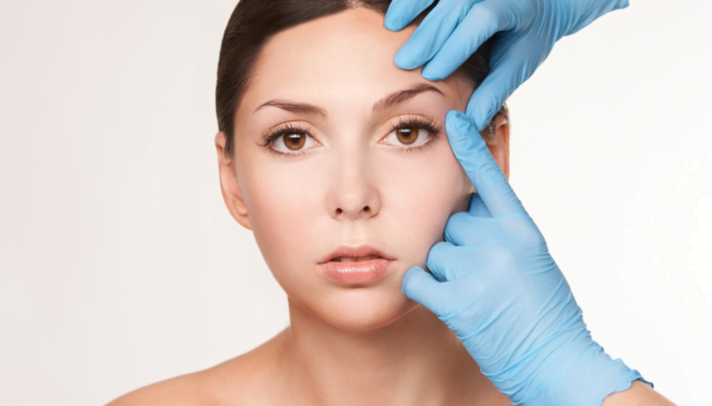 A plastic surgeon in blue gloves examining a woman's face, focusing on the forehead and temple area.