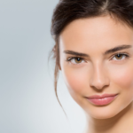 Eine Nahaufnahme einer jungen Frau mit natürlichem Make-up, reiner Haut und zu einem ordentlichen Zopf zurückgeflochtenem Haar vor einem hellgrauen Hintergrund.
