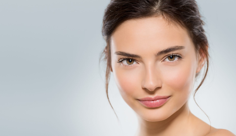 Una imagen de primer plano de una mujer joven con maquillaje natural, piel clara y su cabello recogido en una trenza ordenada, sobre un fondo gris claro.