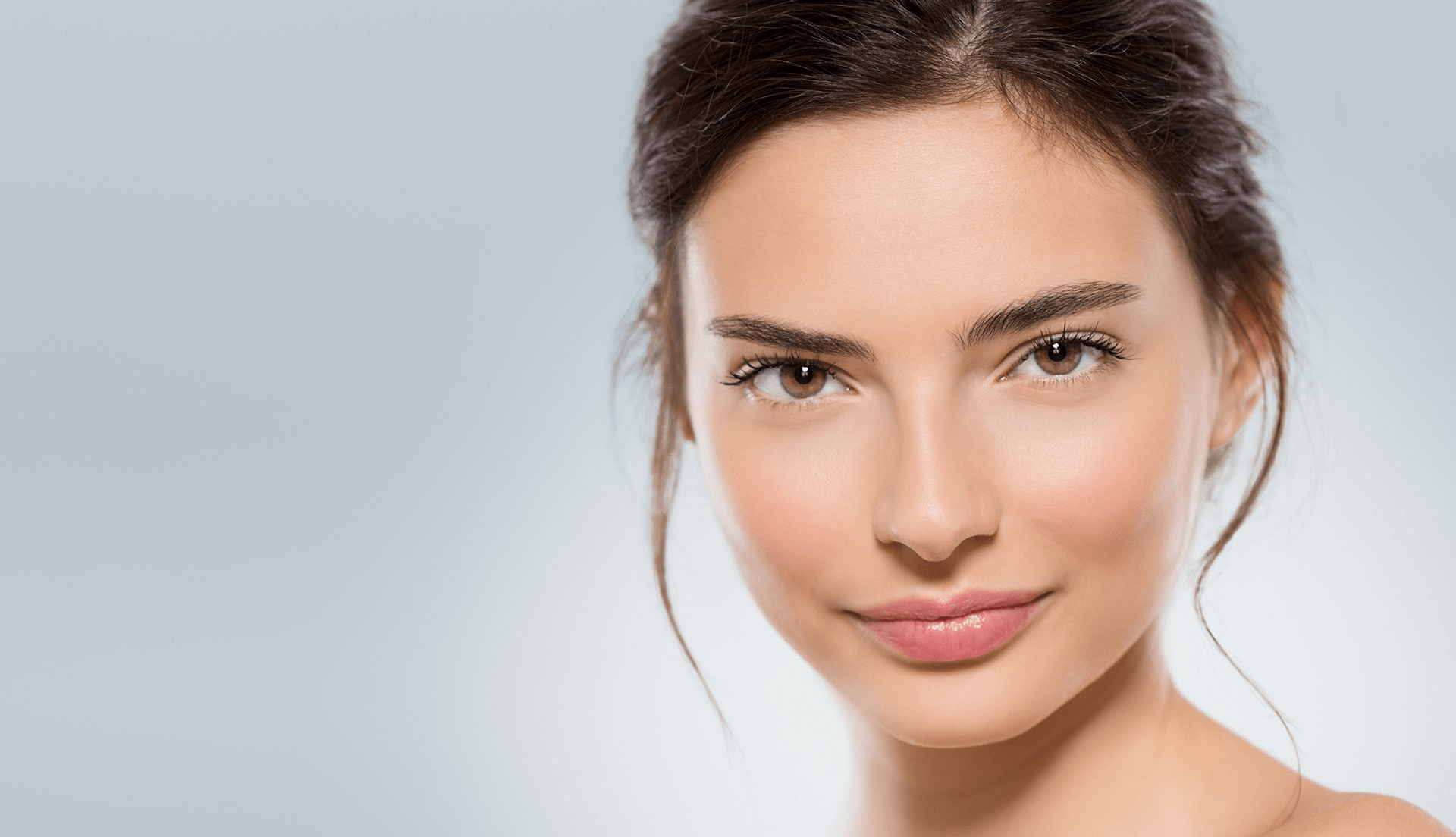 Een close-upfoto van een jonge vrouw met natuurlijke make-up, een heldere huid en haar haar netjes in een vlecht, tegen een lichtgrijze achtergrond.