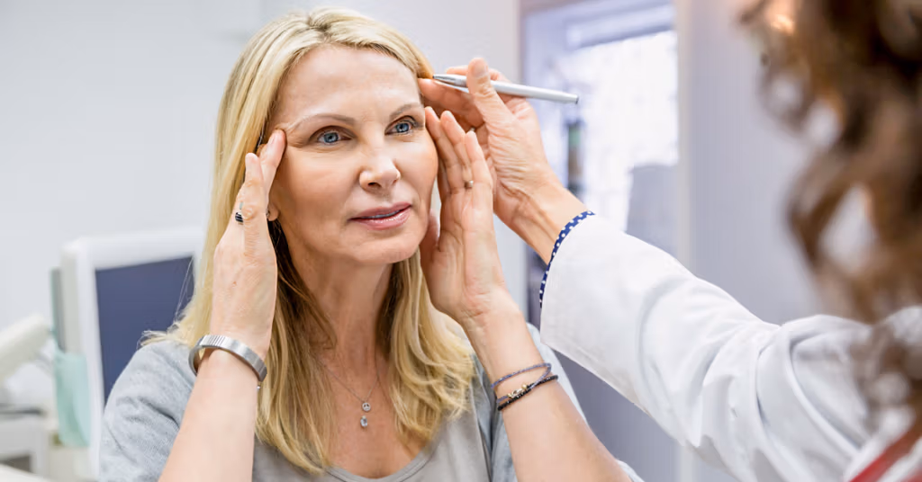 Une maquilleuse professionnelle applique du maquillage à une femme mûre, en se concentrant sur ses sourcils, dans un salon bien éclairé.