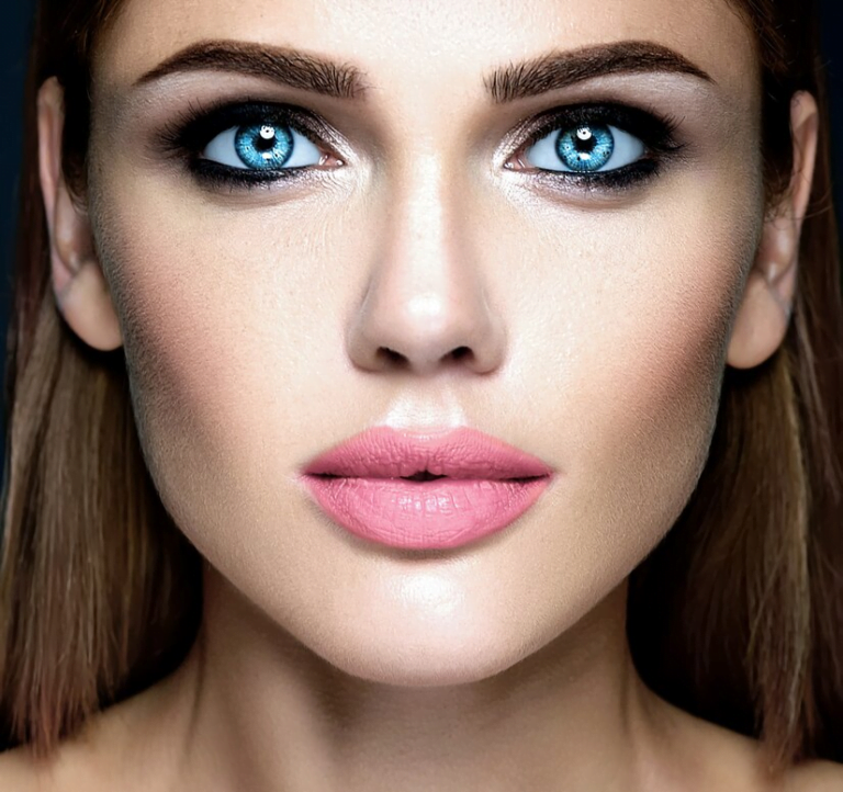 Un retrato en primer plano de una mujer con llamativos ojos azules, maquillaje contorneado y lápiz labial rosa, sobre un fondo oscuro.