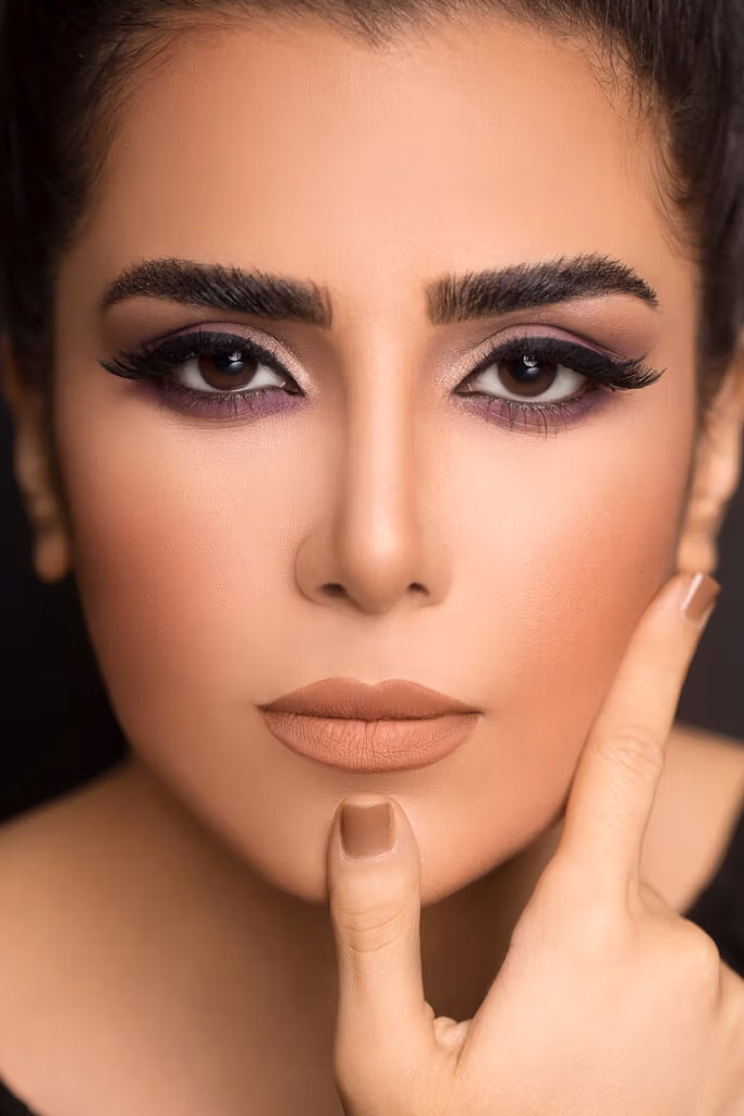 صورة مقربة لامرأة ذات مكياج عيون لافت للنظر وأحمر شفاه بلون البشرة، وهي تتخذ وضعية معينة وتلمس وجهها بأصابعها برفق.