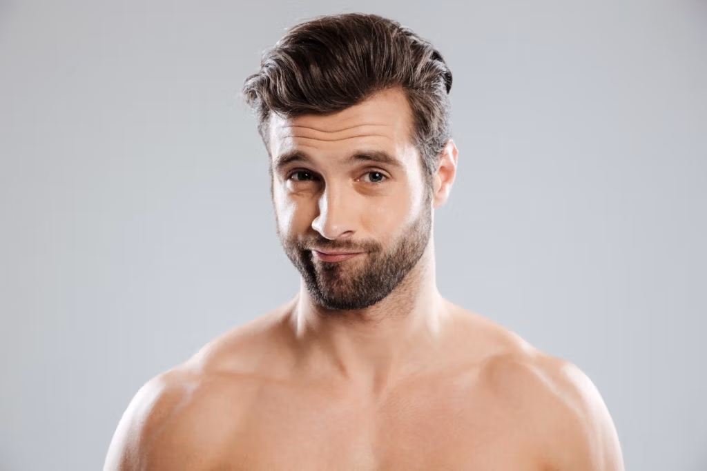 Ein Mann ohne Hemd, mit Bart und kurzen dunklen Haaren, der mit neutralem Gesichtsausdruck direkt in die Kamera blickt.