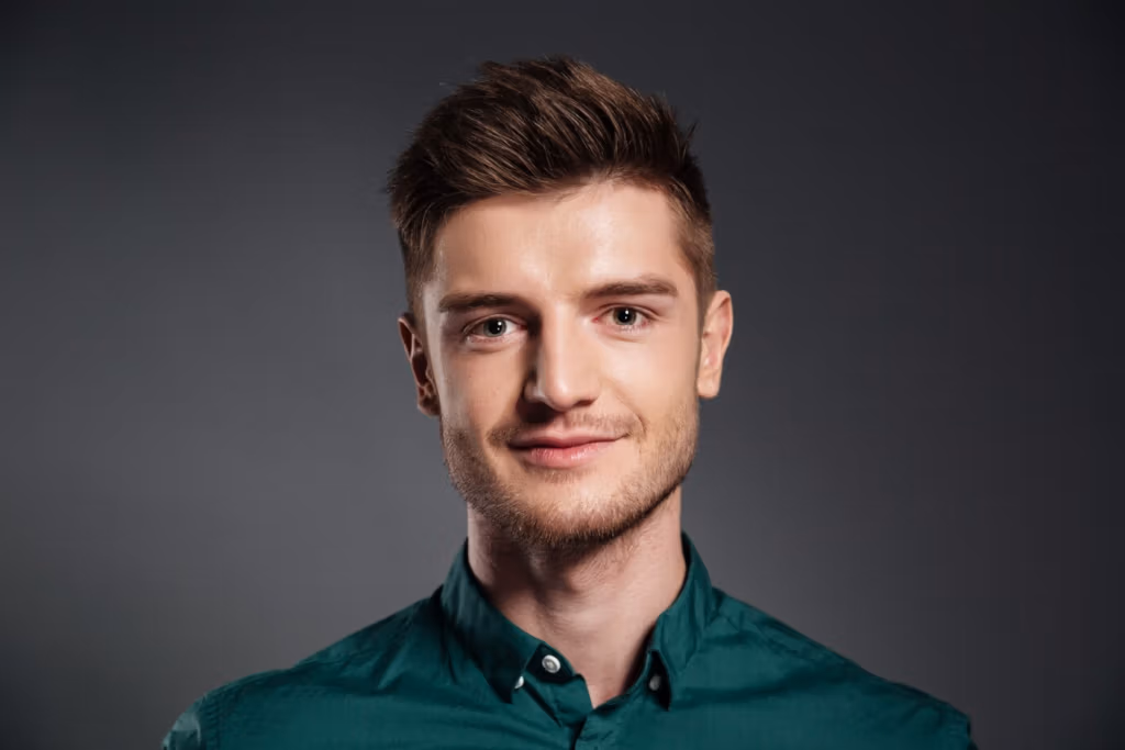 Ein Porträt eines jungen Mannes mit kurzen braunen Haaren, der ein grünes Hemd mit Knöpfen trägt und vor einem dunkelgrauen Hintergrund posiert.