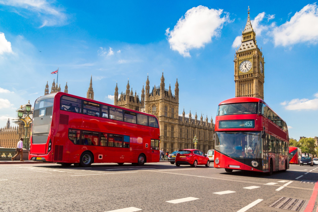Une scène de rue animée à Londres mettant en vedette deux bus rouges emblématiques à impériale passant devant les Chambres du Parlement et Big Ben sous un ciel bleu clair.