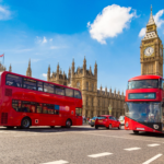 Une scène de rue animée à Londres mettant en vedette deux bus rouges emblématiques à impériale passant devant les Chambres du Parlement et Big Ben sous un ciel bleu clair.