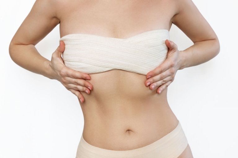 Una mujer que lleva un sujetador sin tirantes de color beige, bajándolo para revelar su pecho, de pie frente a un fondo blanco liso.