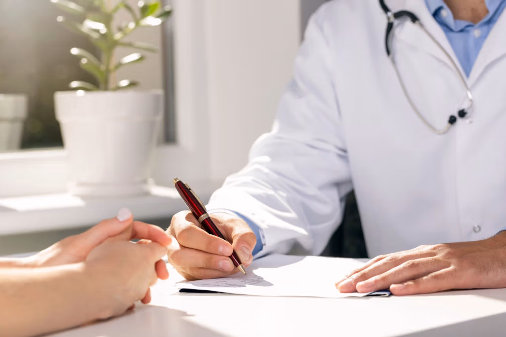 Un médico con bata blanca escribe en un documento con un bolígrafo rojo mientras un paciente está sentado frente a él en un escritorio, con una planta en maceta y una ventana al fondo.