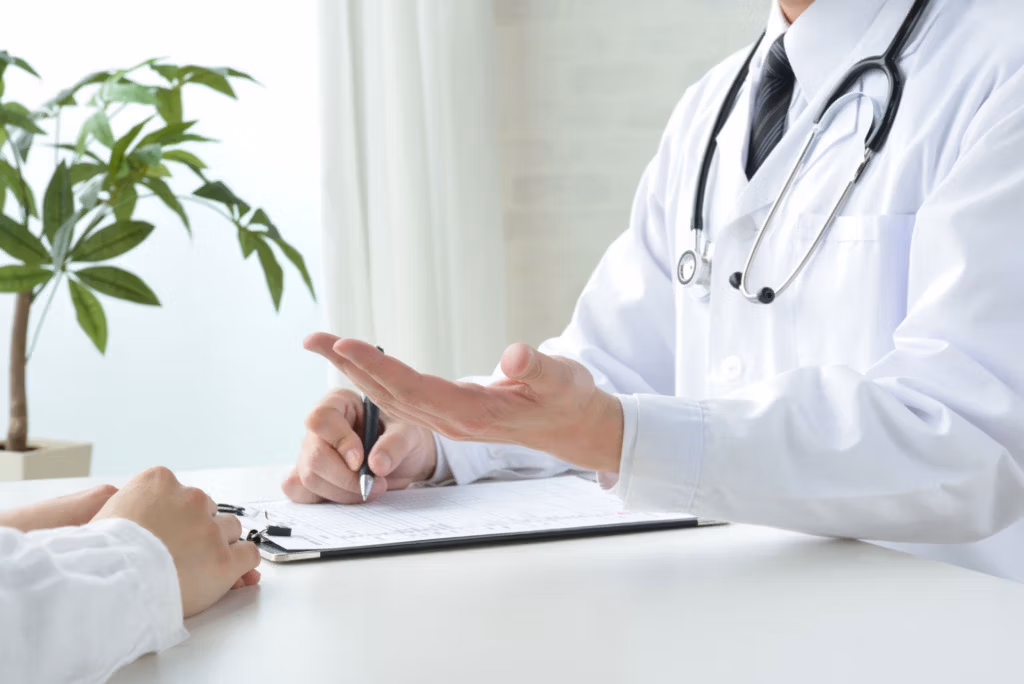 Un médico con bata blanca y un estetoscopio al cuello está sentado en un escritorio, explicando información en un portapapeles a un paciente. La escena se desarrolla en un consultorio médico bien iluminado, con una planta al fondo.