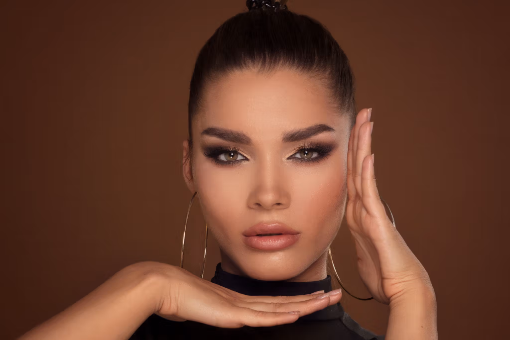 Une femme aux cheveux noirs attachés en chignon, portant de grandes boucles d'oreilles créoles et posant avec ses mains encadrant son visage.