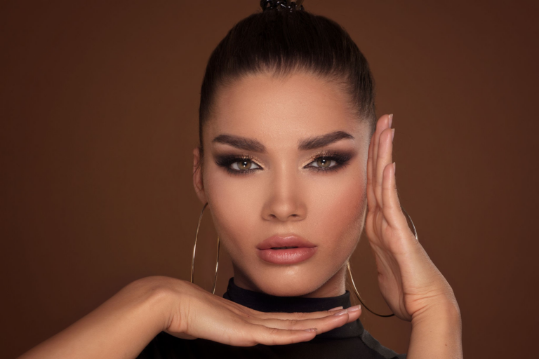 Una mujer con cabello oscuro atado en un moño, con grandes aros y posando con sus manos enmarcando su rostro.