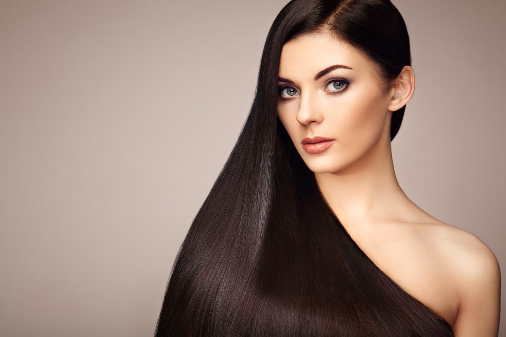 A woman with long, straight, dark hair draped over her shoulder, gazing directly at the camera with a neutral expression. The background is plain and light-colored, allowing her hair and facial features to be the focal point.