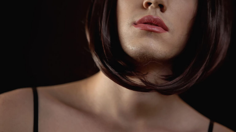 Primer plano del rostro de una mujer, con énfasis en los labios y la barbilla, con piel suave e iluminación sutil. El fondo es oscuro, lo que realza los rasgos faciales. La mujer tiene cabello oscuro hasta los hombros que le cubre parcialmente el cuello y los hombros.