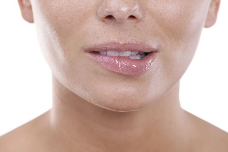 Primer plano de la parte inferior del rostro de una persona, con énfasis en los labios y la barbilla. Los labios se ven húmedos y ligeramente brillantes, mientras que la piel de la barbilla y la zona circundante es lisa, con algunos poros visibles. El fondo es liso y blanco, lo que realza los rasgos faciales.